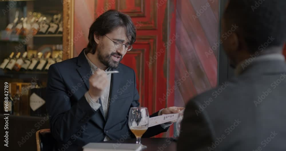 Middle Eastern businessman talking to African American partner pointing at document then clinking glasses celebrating partnership in cafe. Teamwork and informal meeting concept. Middle Eastern businessman talking to African American partner pointing at document then clinking glasses celebrating partnership in cafe. Teamwork and informal meeting concept.