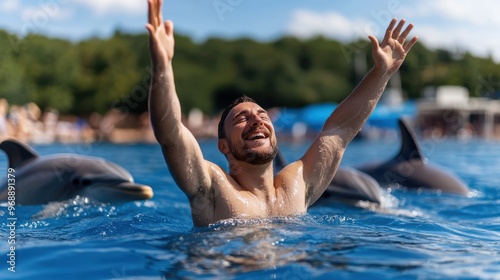 A person in water, lifting arms upward amid two dolphins, showcases an energetic and joyful interaction with nature, capturing a moment of playful exuberance under a sunny sky. photo