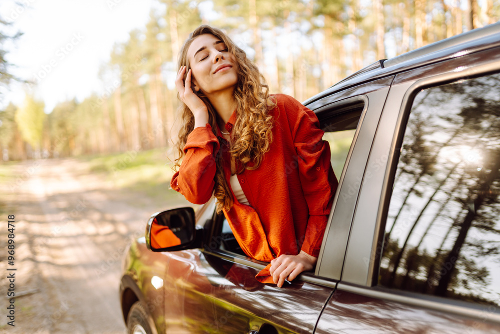 Naklejka premium The young woman leans out of the car window, basking in the sunlight and enjoying the natural surroundings of a serene forest road. The concept of active lifestyle, travel, tourism.