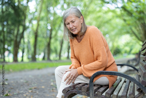 Senior woman feeling knee pain sitting on bench in park, showing discomfort. Mature adult dealing with joint pain outdoors in tranquil environment, highlighting aging and health challenges. photo