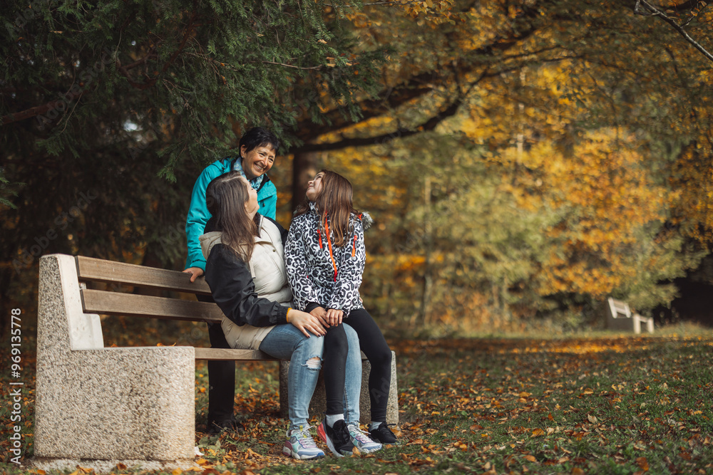 Fototapeta premium A joyful family moment in a park during autumn. Three women are sitting on a bench, laughing and enjoying each other's company amidst colorful fall foliage.