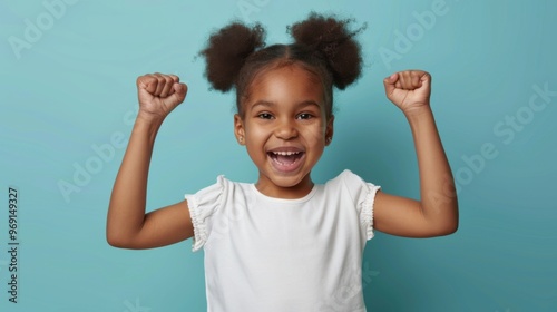 A little Black girl with a big smile and her fists raised in the air celebrates a victory or achievement. She is wearing a white shirt and her dark hair is styled in a high ponytail, copy space photo