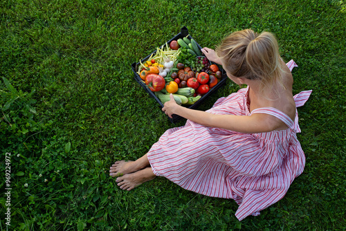 Top view of a basket of fresh vegetables standing on green grass. The vegetables are ripe and juicy. A blonde woman in a striped dress sits next to the basket and holds a plastic basket in her hands