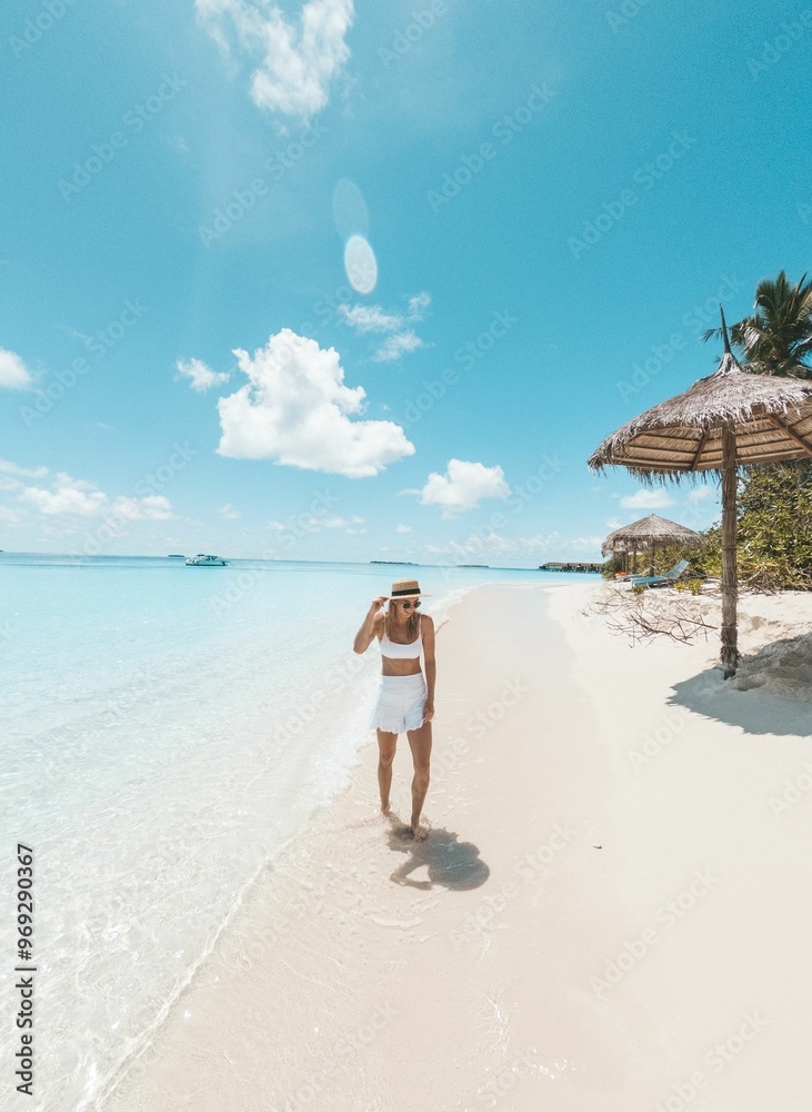 Obraz premium Woman walking along a pristine beach with clear blue water and a bright sky.