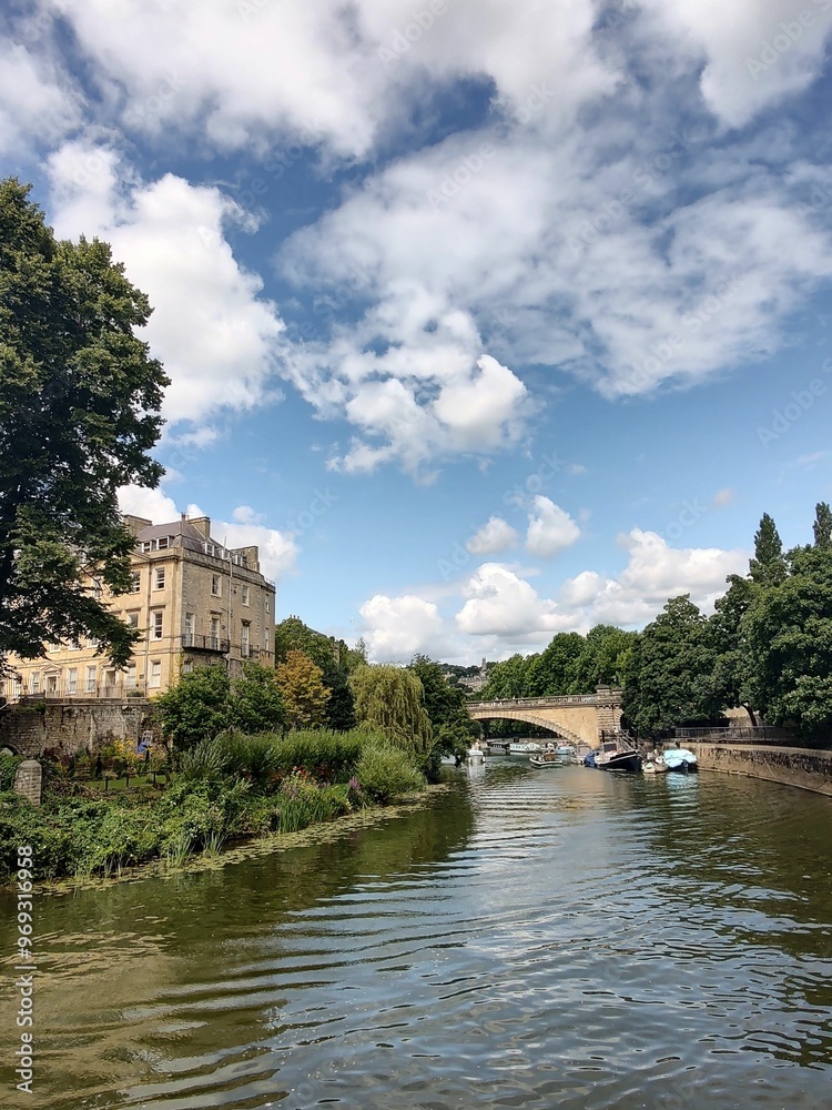 A river with boats, lush greenery, and a stone bridge under a partly cloudy sky in Bath A river with boats, lush greenery, and a stone bridge under a partly cloudy sky in Bath