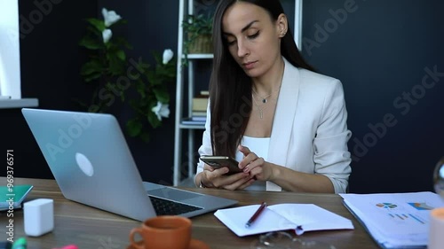 Businesswoman in light blazer multitasks at her desk, talking on the phone while typing on a laptop. The workspace is tidy, featuring documents, and a notebook, creating a professional atmosphere