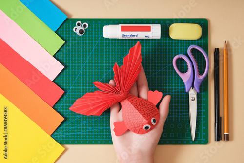 Women's hands hold a craft red paper fish against a green paper craft mat. Scissors, colored paper, glue, felt-tip pen. Top view