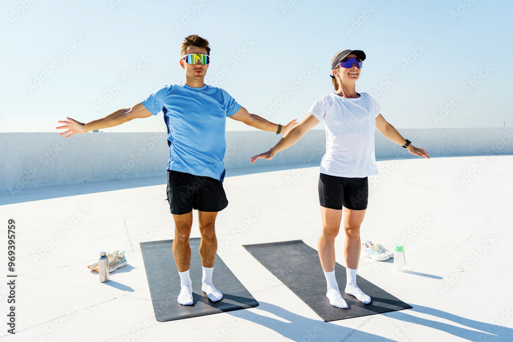 A couple stands on yoga mats, arms outstretched in a morning stretch routine under a clear sky. A couple stands on yoga mats, arms outstretched in a morning stretch routine under a clear sky.
