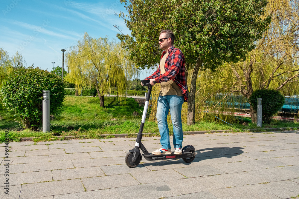 Side view of man riding e-scooter on pavement surface in city public park at sunny summer day. Male driving electric scooter outdoors. Eco-friendly city transport and travel. Sustainable lifestyle Side view of man riding e-scooter on pavement surface in city public park at sunny summer day. Male driving electric scooter outdoors. Eco-friendly city transport and travel. Sustainable lifestyle