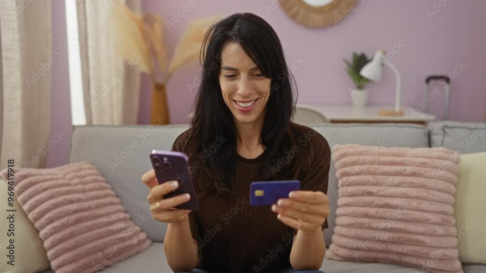 Woman using smartphone holding credit card sitting on cozy sofa in living room with decorative pillows and indoor plants Woman using smartphone holding credit card sitting on cozy sofa in living room with decorative pillows and indoor plants