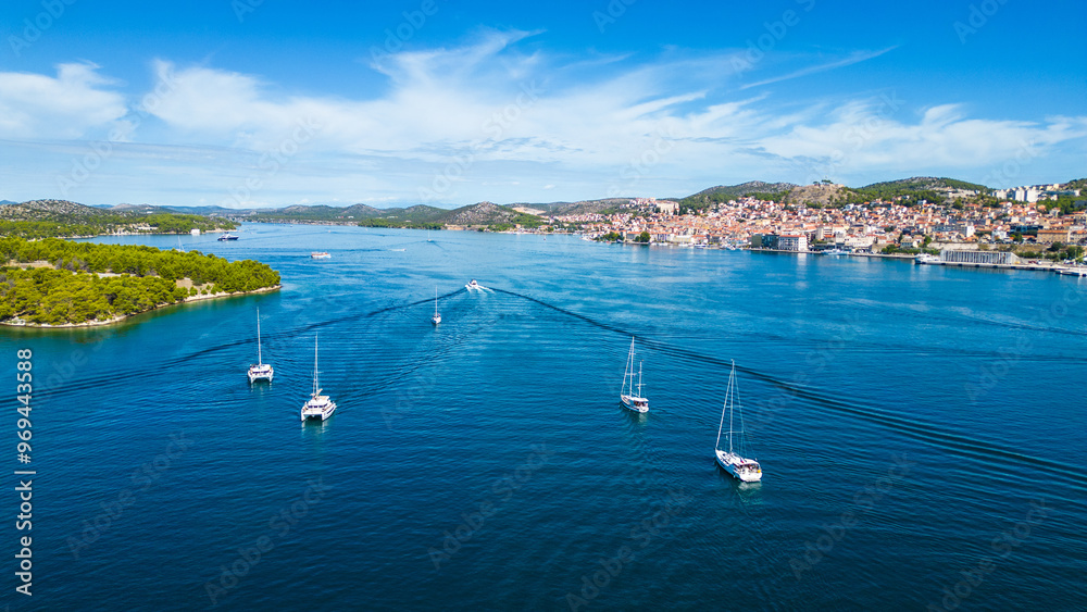 Obraz premium A yacht departs from the marina in Šibenik, Croatia, gliding through the calm waters of the Adriatic Sea. This aerial view captures the scenic coastline, with the historic city of Šibenik 