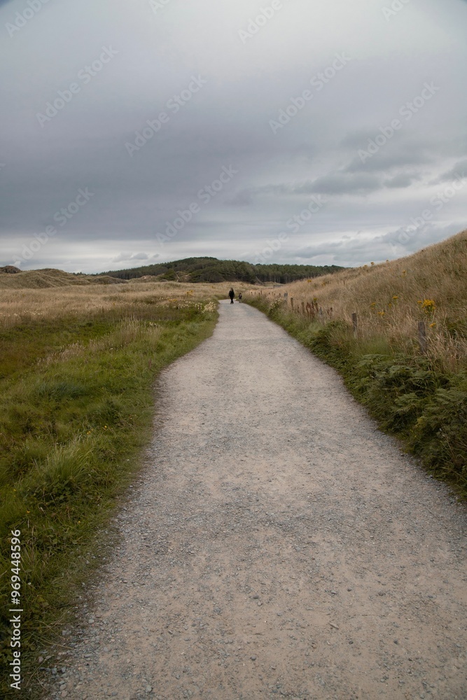 Solitary person walking along a gravel path through a grassy landscape under a cloudy sky Solitary person walking along a gravel path through a grassy landscape under a cloudy sky