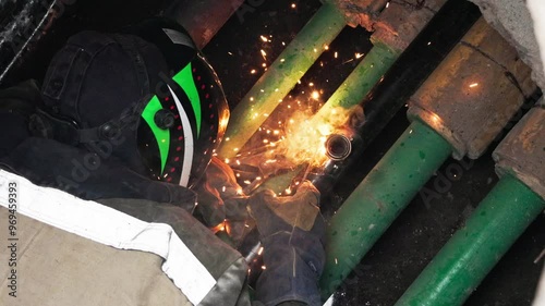 A welder donned in protective gear welds green pipes, producing bright sparks in a dim industrial space, highlighting the meticulous nature and intensity of this labor-intensive task. photo