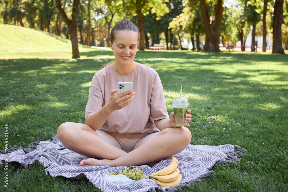 Pretty pleased young woman relaxing in summer attire on grass using her phone to scroll online and enjoy the sunny day at picnic chatting online with friends Pretty pleased young woman relaxing in summer attire on grass using her phone to scroll online and enjoy the sunny day at picnic chatting online with friends