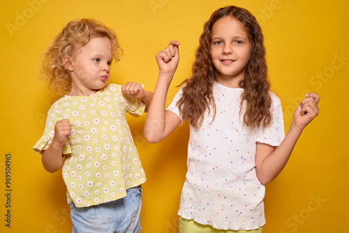 Blonde toddler and brown-haired sibling dancing joyfully and celebrating with smiles and excitement expressing victory and happiness while standing indoors isolated on yellow background photo