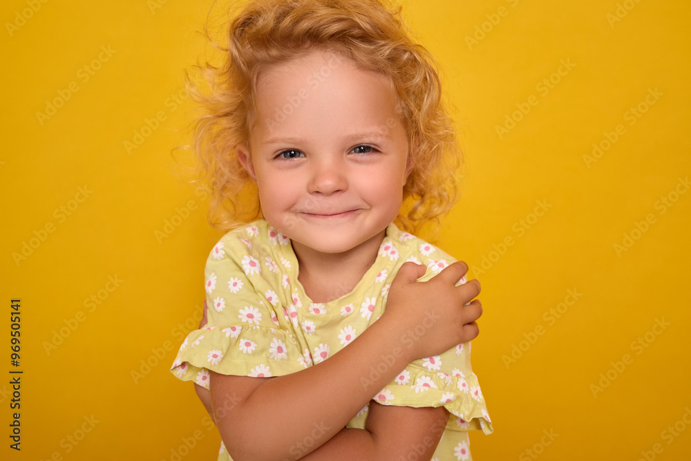 Blonde curly-haired Caucasian toddler girl with satisfied expression and charming smile embracing herself while standing alone indoors on yellow background Blonde curly-haired Caucasian toddler girl with satisfied expression and charming smile embracing herself while standing alone indoors on yellow background