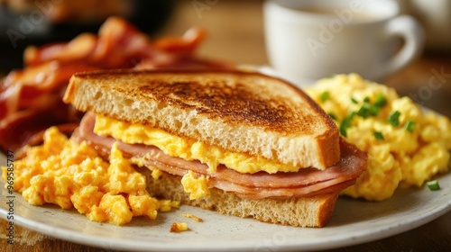 A breakfast spread featuring a toasted ham and cheese sandwich with scrambled eggs and a side of bacon.