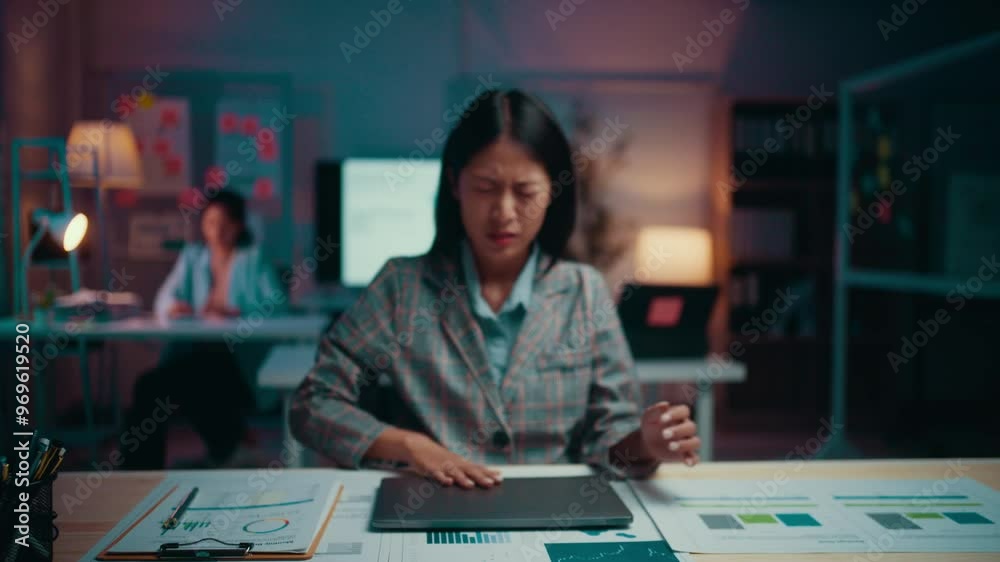 Asian businesswoman is working late in the office. She is getting increasingly frustrated with her work. Her colleague is working in the background Asian businesswoman is working late in the office. She is getting increasingly frustrated with her work. Her colleague is working in the background