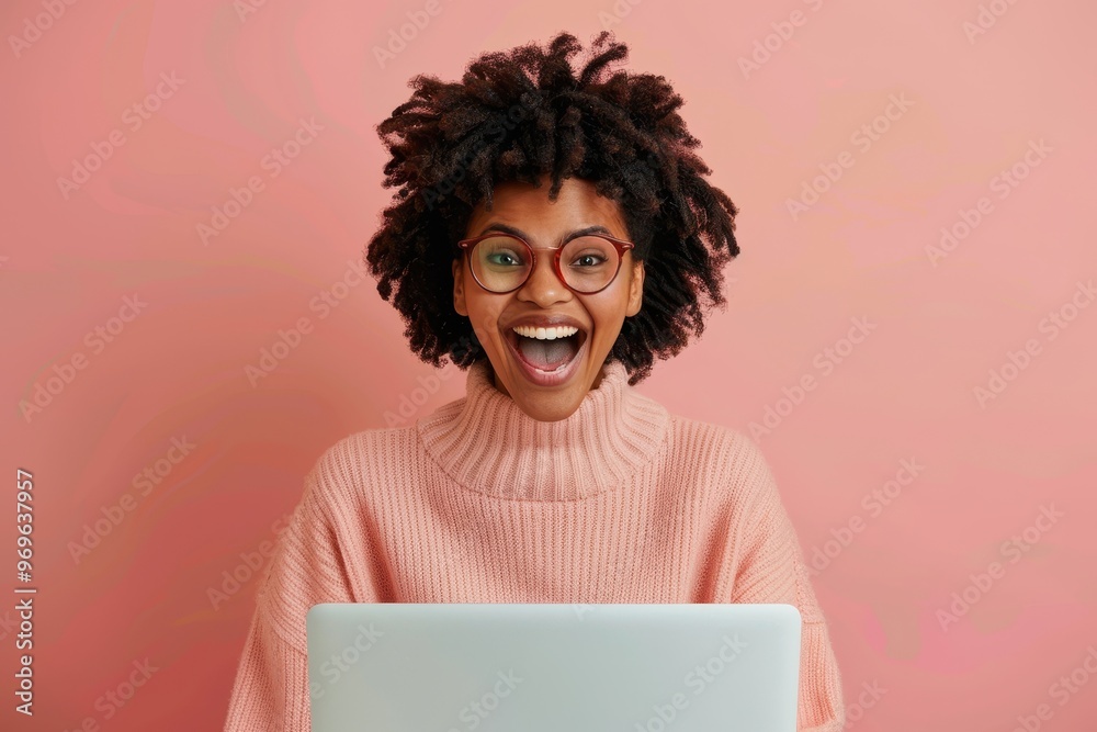 Cheerful young woman with an afro hairstyle and glasses, wearing a pink sweater, expressing excitement while using a laptop, set against a pink background. Cheerful young woman with an afro hairstyle and glasses, wearing a pink sweater, expressing excitement while using a laptop, set against a pink background.