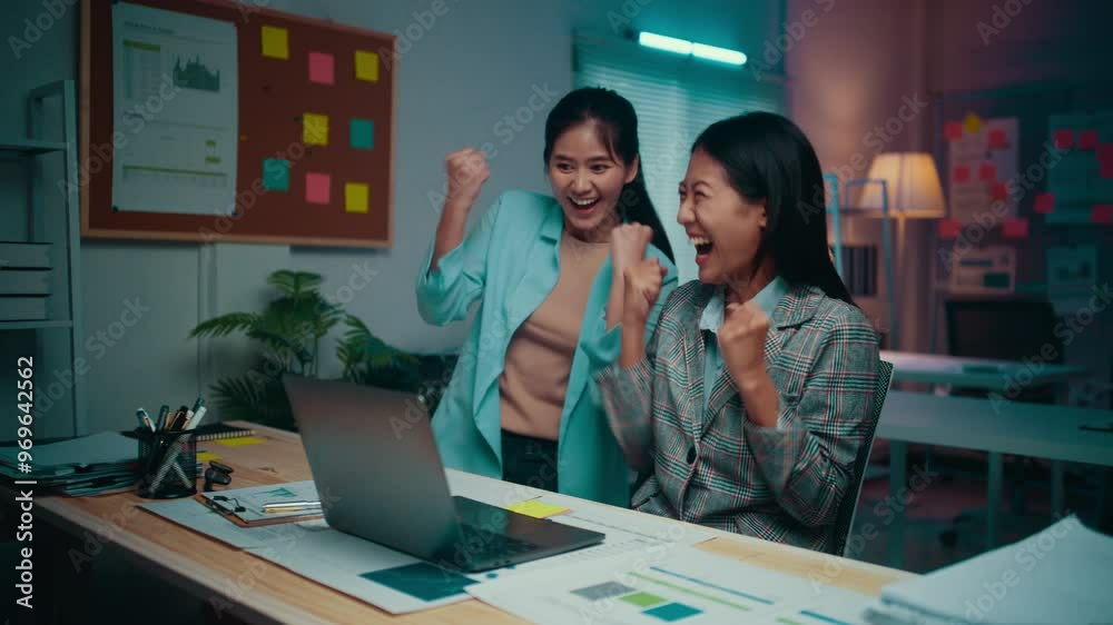 Excitement fills the office as two young businesswomen celebrate a significant achievement while working on a laptop at night, showcasing their teamwork and success Excitement fills the office as two young businesswomen celebrate a significant achievement while working on a laptop at night, showcasing their teamwork and success