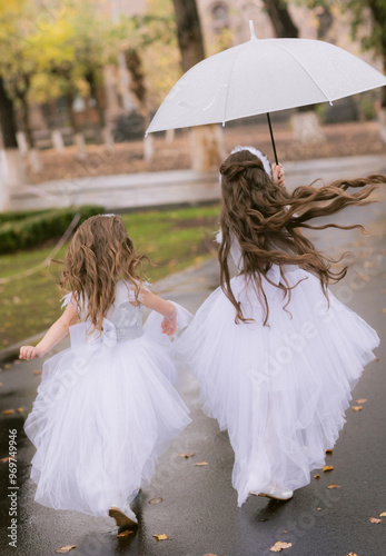 Backshot of two girls in long white celebration dresses running along a street with umbrella photo