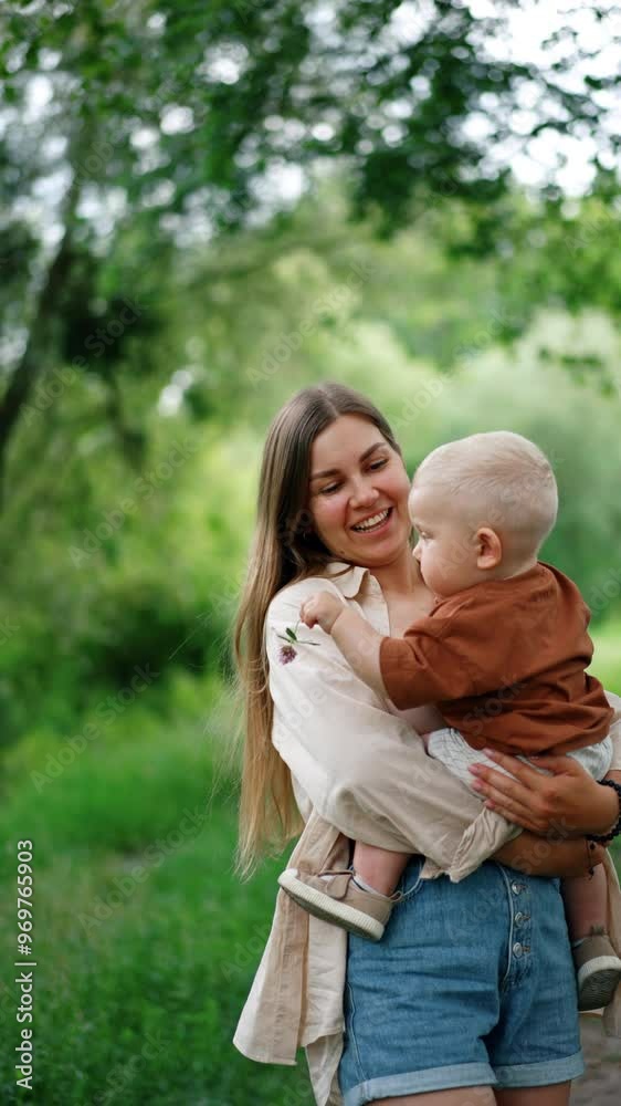 Long-haired Caucasian woman holding her baby in hands. Kid with flower in his hands looks around at nature with interest. Vertical video. Long-haired Caucasian woman holding her baby in hands. Kid with flower in his hands looks around at nature with interest. Vertical video.