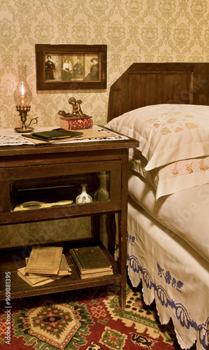A vintage bedroom with wooden bed, pillows, embroidered pillowcases and valance, bedside table with books, perfume and lamp with burning candle, and vintage photographs (blurred for copyright purpose)