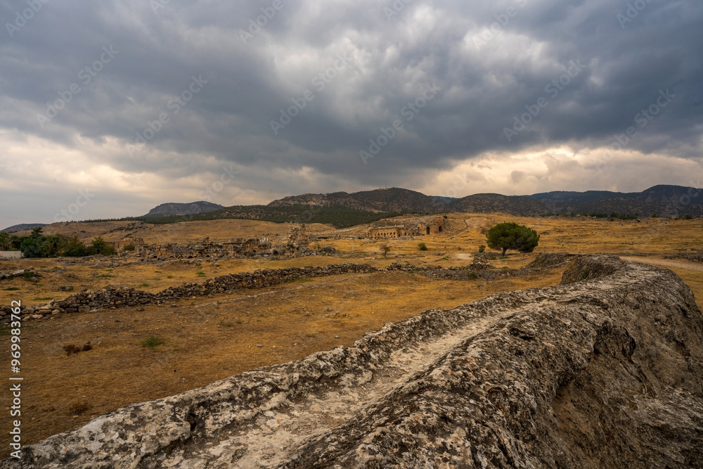 Hierapolis in Pamukkale: A desolate path winds through the arid landscape, leading towards ancient ruins nestled beneath a dramatic, cloud-filled sky.