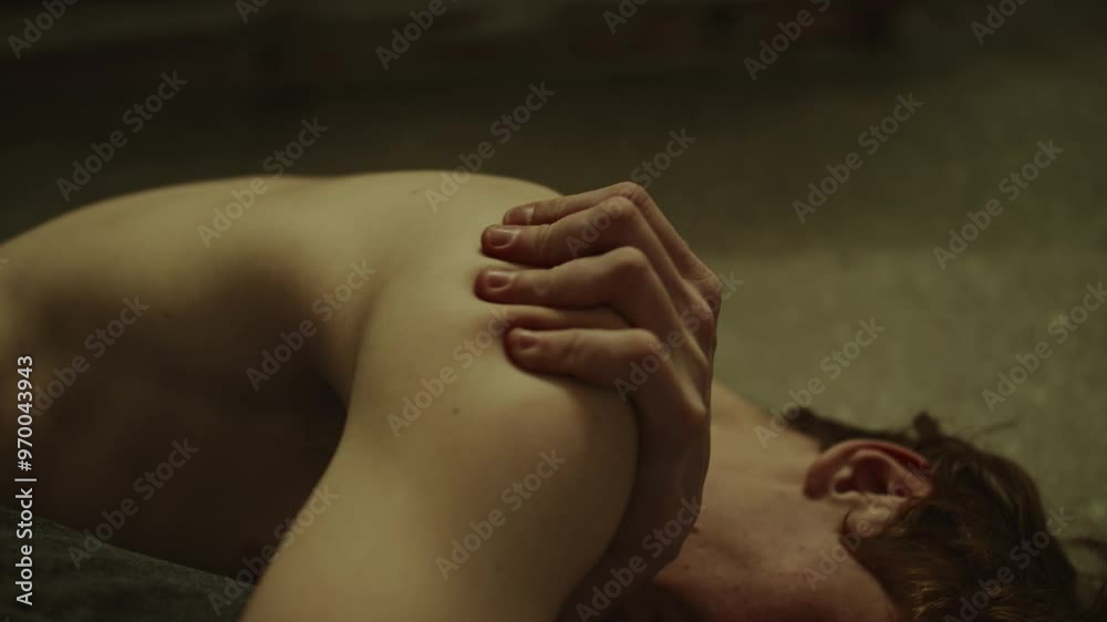 Close-up view of shirtless young man touching shoulder and crying while lying on floor with drawn up knees in fetal position