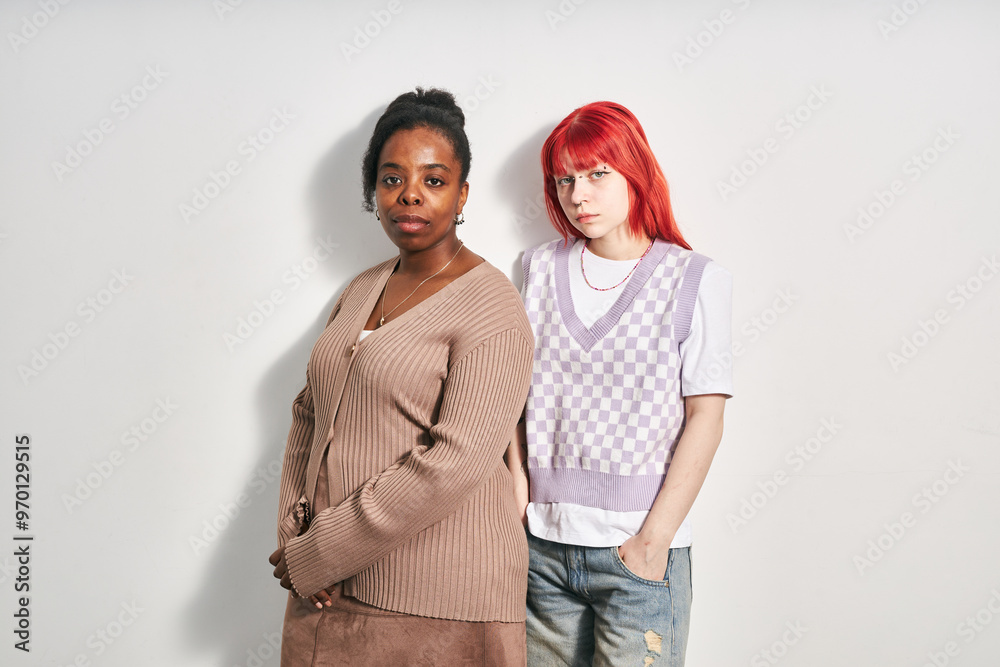 Portrait of two women standing against white background, one with African descent wearing a beige sweater, and another with red hair in casual clothing, looking at camera Portrait of two women standing against white background, one with African descent wearing a beige sweater, and another with red hair in casual clothing, looking at camera
