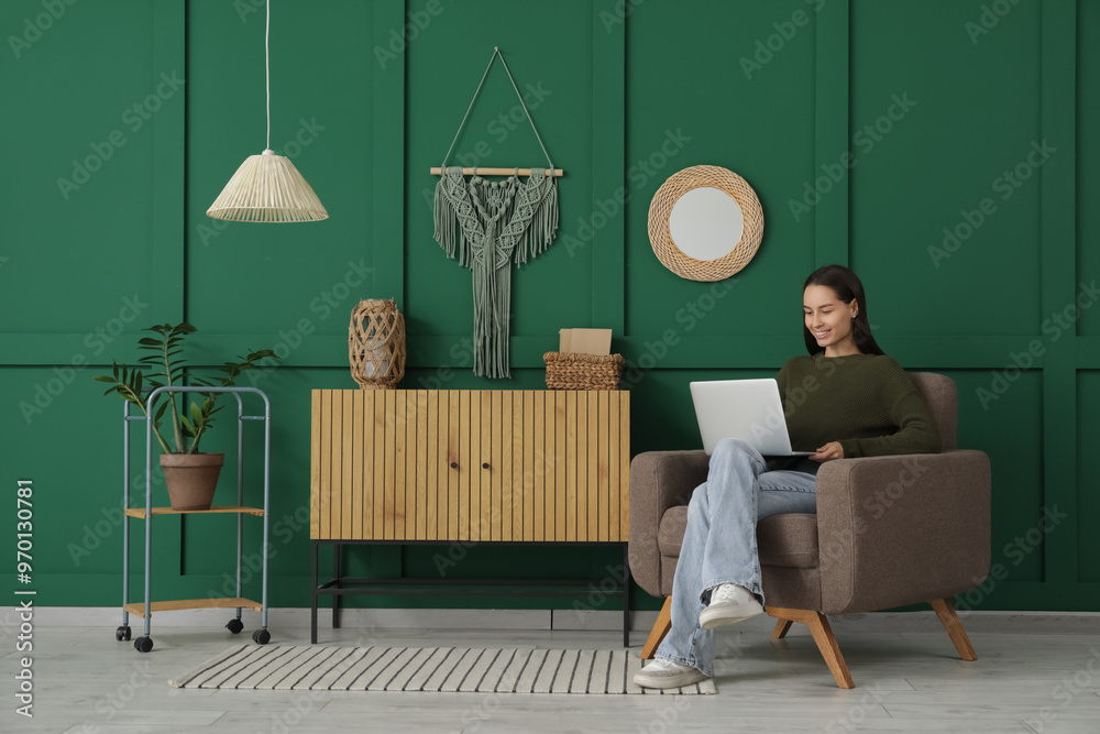 Fototapeta premium Young woman using laptop while sitting in armchair at home