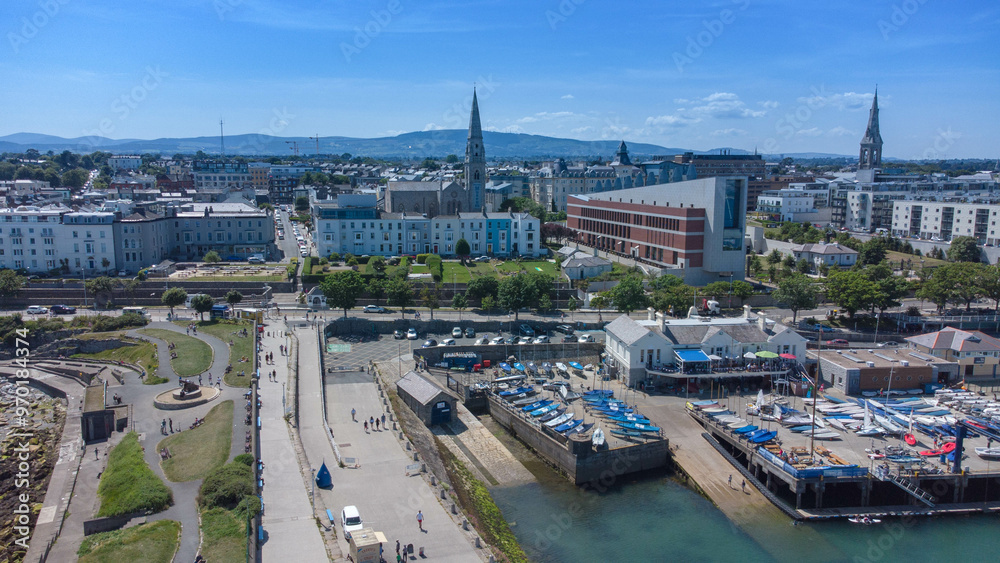 Fototapeta premium An aerial drone shot of Dun Laoghaire, Ireland, captures the picturesque coastal town from above. The image showcases the bustling harbor, with boats and ships anchored in the clear blue waters.