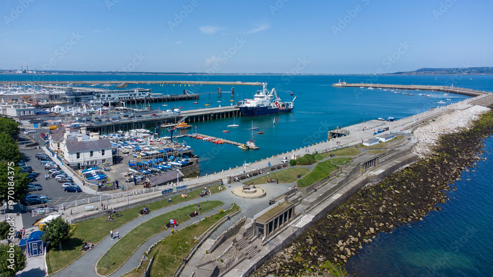 Fototapeta premium An aerial drone shot of Dun Laoghaire, Ireland, captures the picturesque coastal town from above. The image showcases the bustling harbor, with boats and ships anchored in the clear blue waters.