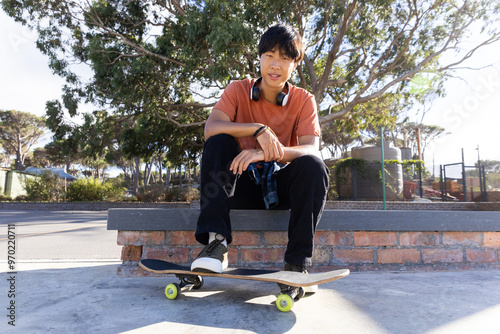 Sitting on skateboard, asian teenage boy with headphones relaxing outdoors in park