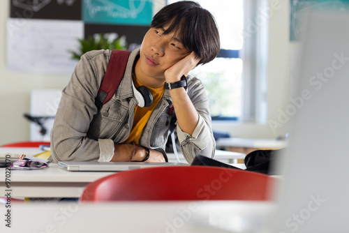 In school, asian teenage boy sitting at desk looking bored with headphones around neck photo