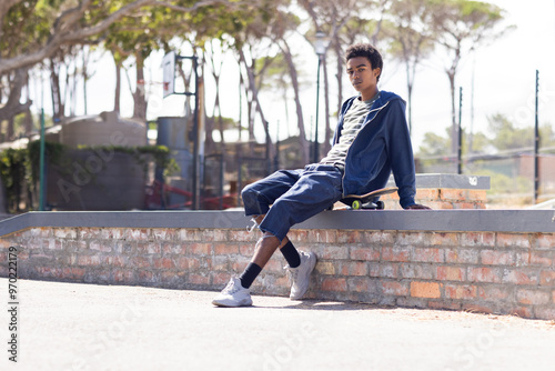 Relaxing outdoors, african american teenage boy sitting on ledge with skateboard nearby