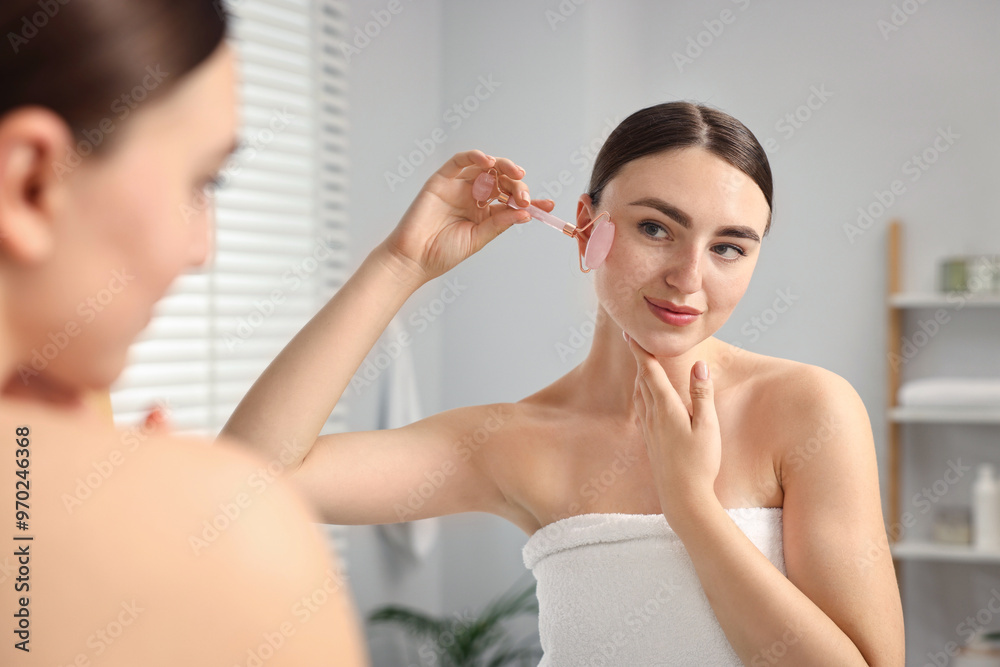 Beautiful young woman doing facial massage with roller near mirror at home