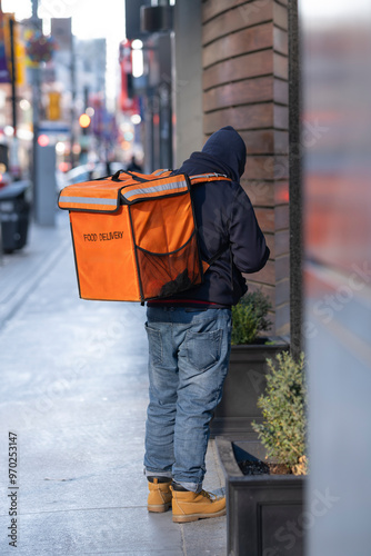 Food courier driver with food delivery sign on the cooler backpack waiting in front of a restaurant for order pick up.