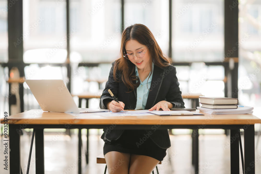 Focused and Determined: A young Asian businesswoman works diligently at her desk, surrounded by paperwork, a laptop, and books. Her focused gaze and determined expression highlight her dedication to s Focused and Determined: A young Asian businesswoman works diligently at her desk, surrounded by paperwork, a laptop, and books. Her focused gaze and determined expression highlight her dedication to s