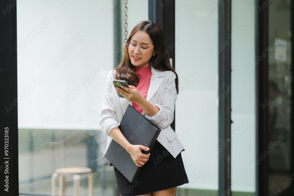 © Kritdanai - Young Professional Navigating the City: A stylish young Asian businesswoman, radiating confidence, checks her phone while carrying a laptop and documents, embodying the modern urban professional lifes