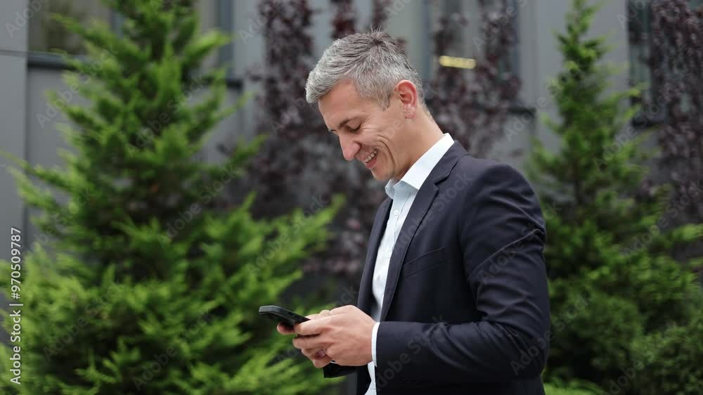 A happy and confident mature businessman in a suit standing outside the office using a smartphone. He smiles as he reads a text message or email. A happy and confident mature businessman in a suit standing outside the office using a smartphone. He smiles as he reads a text message or email.