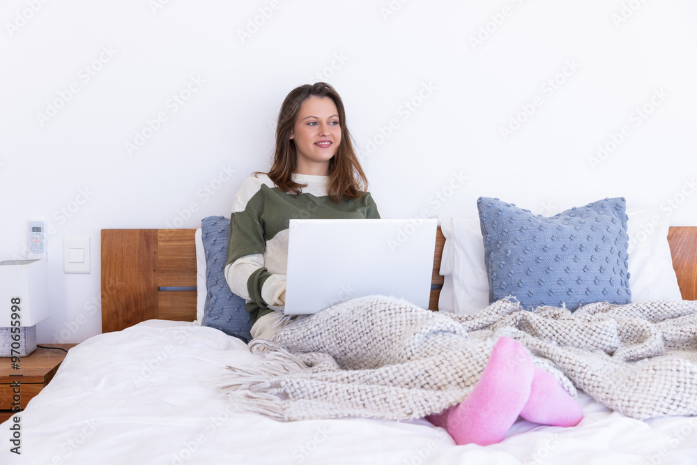 At home, Using laptop, woman relaxing on bed with cozy blanket and pillows At home, Using laptop, woman relaxing on bed with cozy blanket and pillows