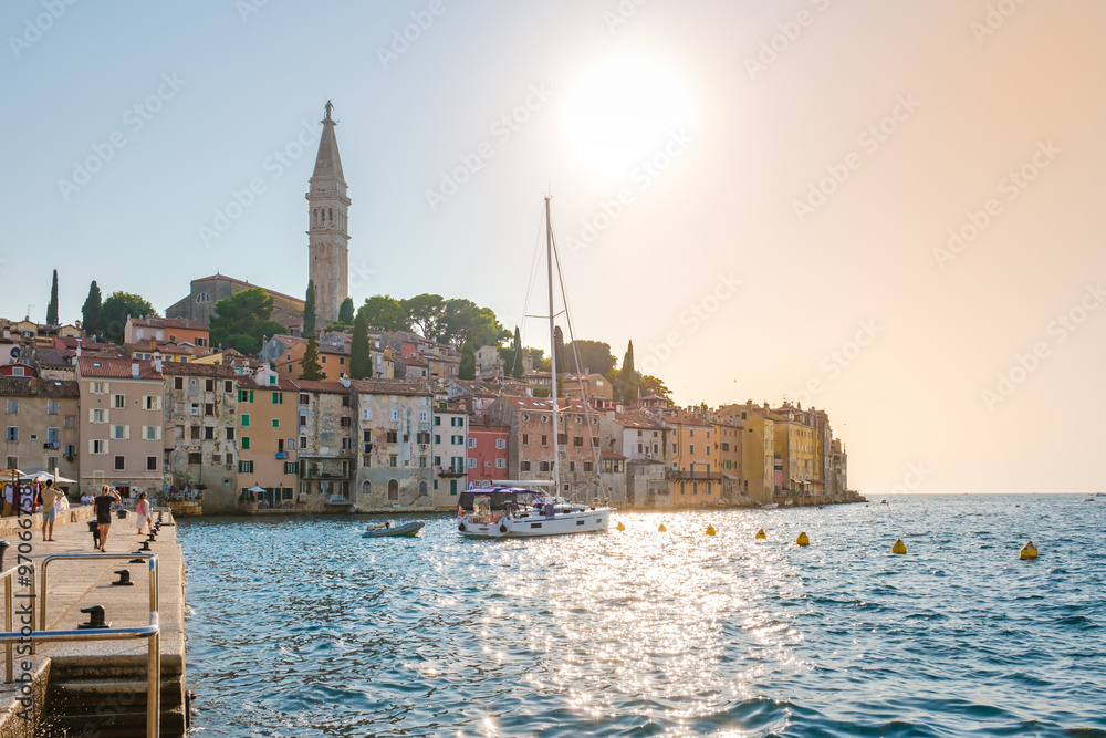 Fototapeta premium The sun casts a warm glow over Rovinj Croatia, illuminating colorful buildings along the waterfront. A sailboat gently floats on the clear waters, inviting exploration of this enchanting coastal town.
