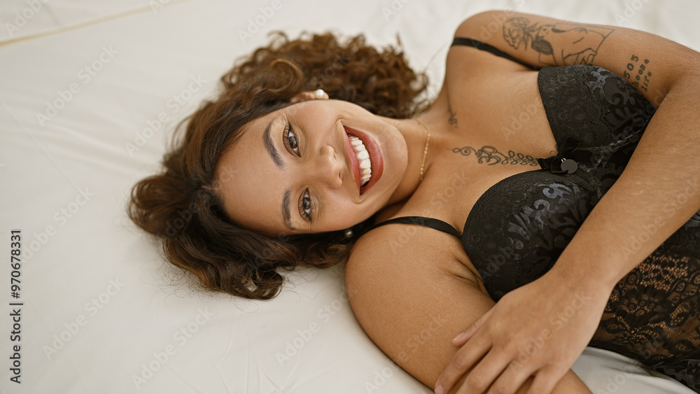 Obraz premium Smiling mixed-race woman lounging in black lingerie on a bedroom's white bedsheet, portraying casual elegance indoors.