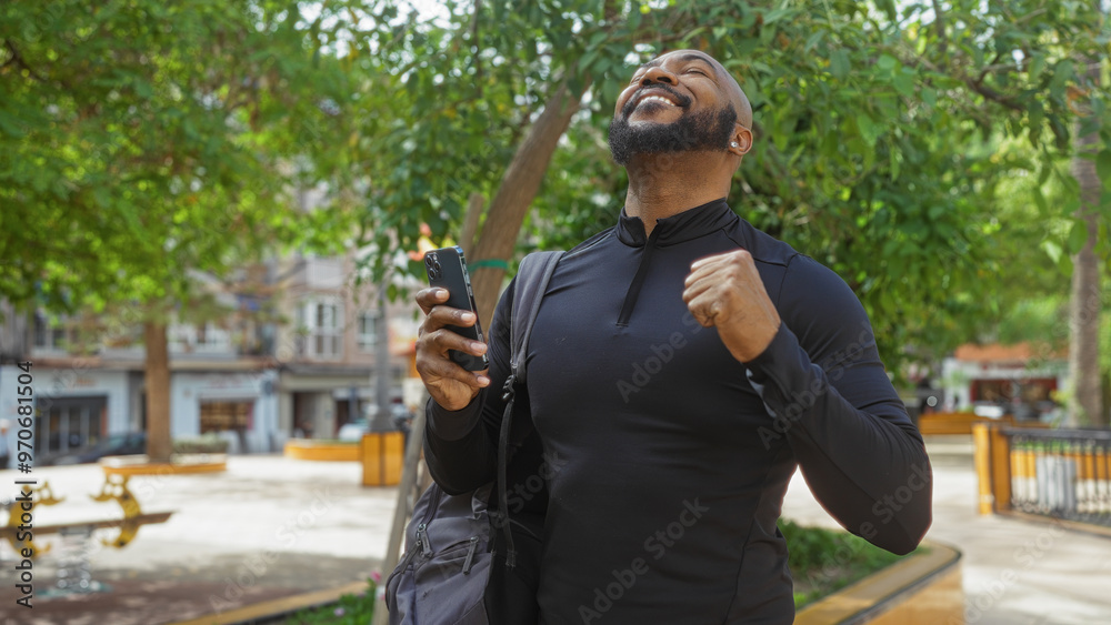 Fototapeta premium Handsome african american man celebrating outdoors in urban park, holding phone and backpack, smiling under trees, feeling victorious