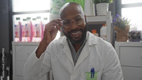 Handsome young man with glasses and beard smiling in a pharmacy wearing a white coat