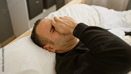 A stressed hispanic man lying on a bed indoors, looking troubled as he holds his forehead, portraying despair or headache.