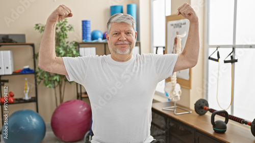 Middle-aged man flexing muscles confidently in a well-equipped physiotherapy clinic.