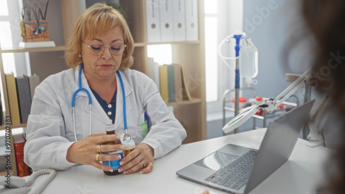 A blonde female doctor in a medical clinic examines a bottle of medication at her desk, surrounded by medical equipment and office supplies.