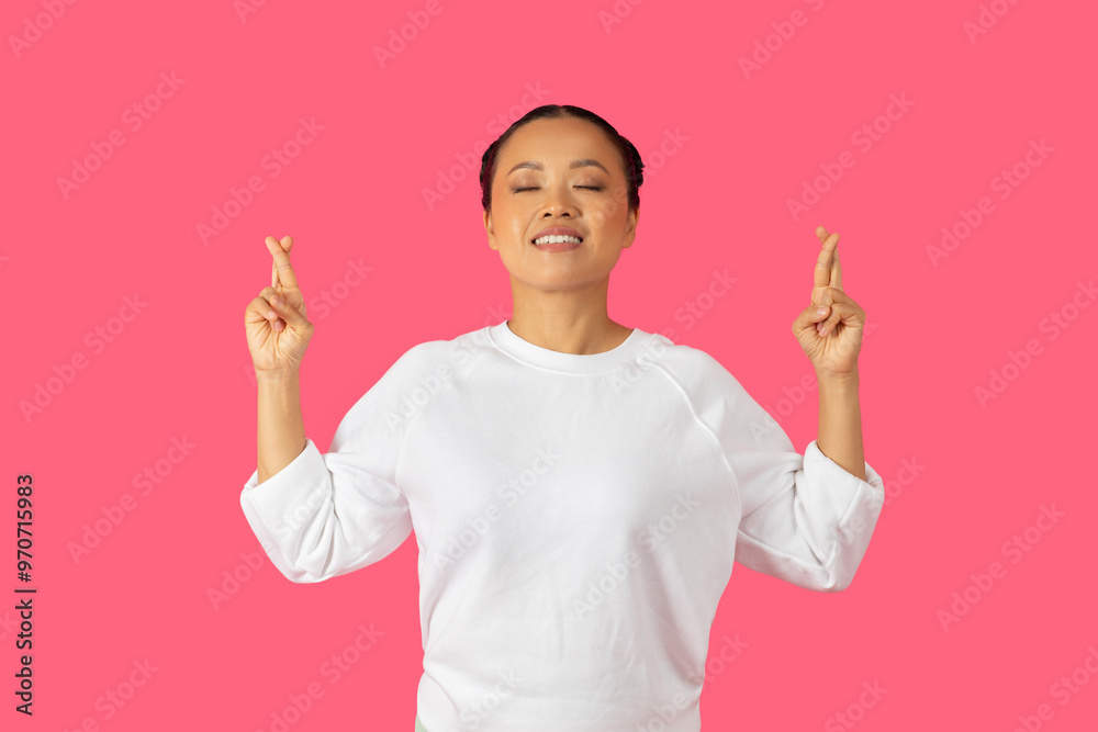 Asian woman expresses hope and positivity with her fingers crossed while smiling against a vibrant pink backdrop.